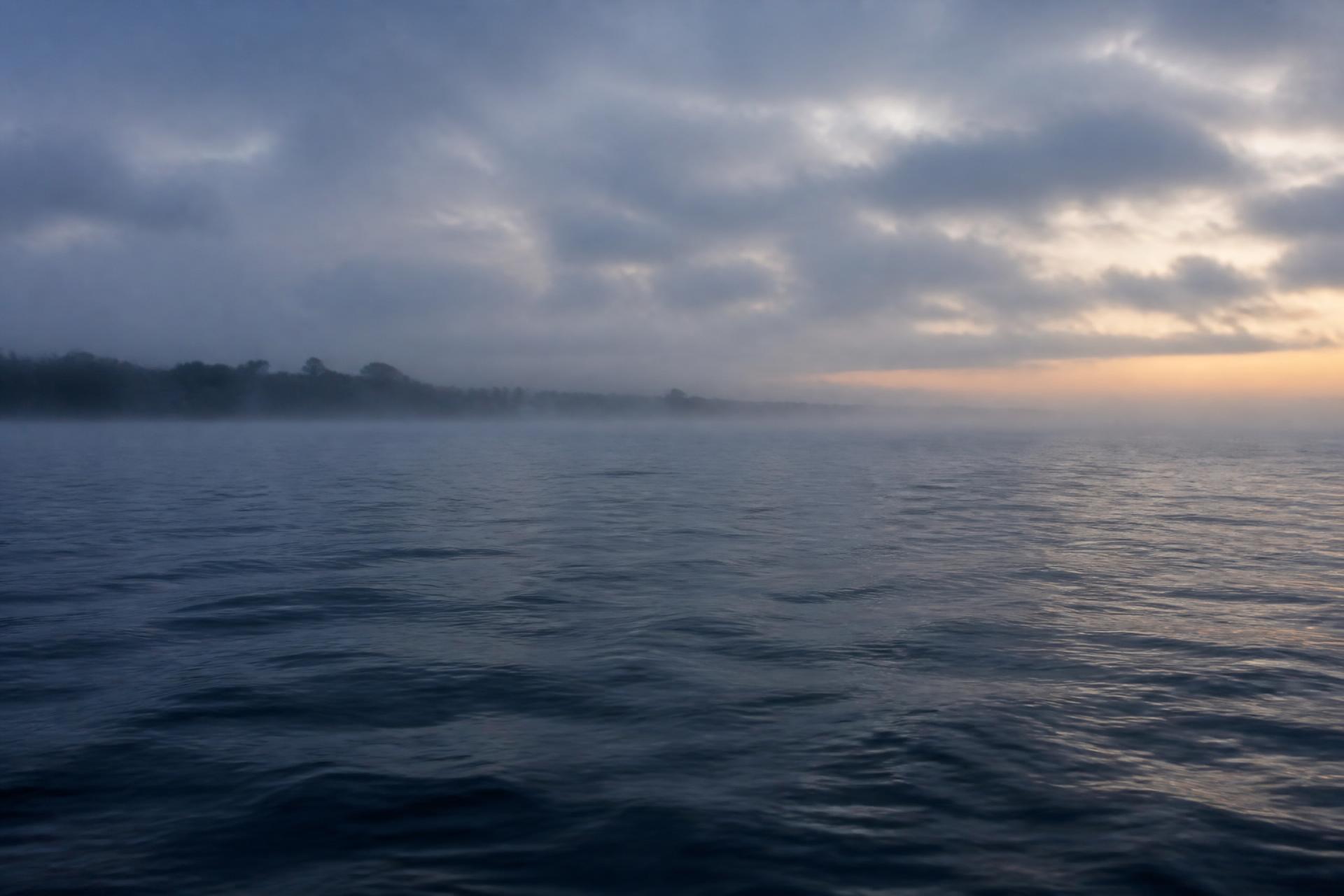 Lake Ontario at dawn, mist over deep blue water near a low forested shoreline